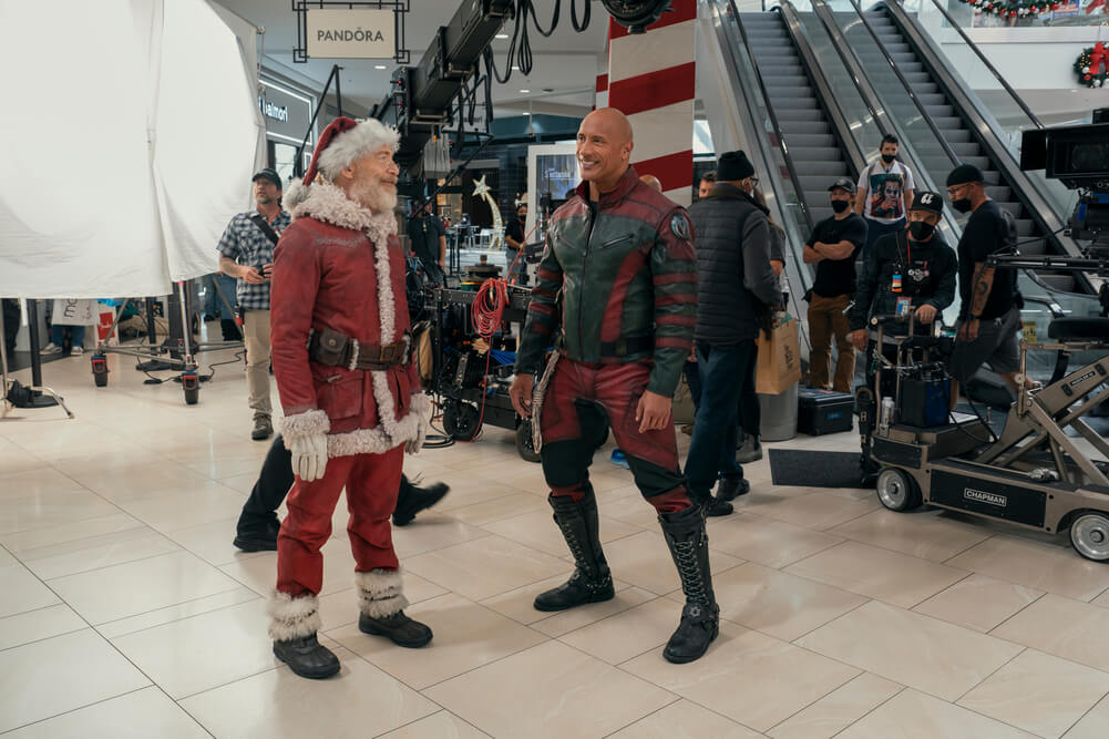 Actor J.K. Simmons stands in a shopping mall dressed as Santa Claus conversing with co-star Dwayne Johnson who plays agent Callum Drift 